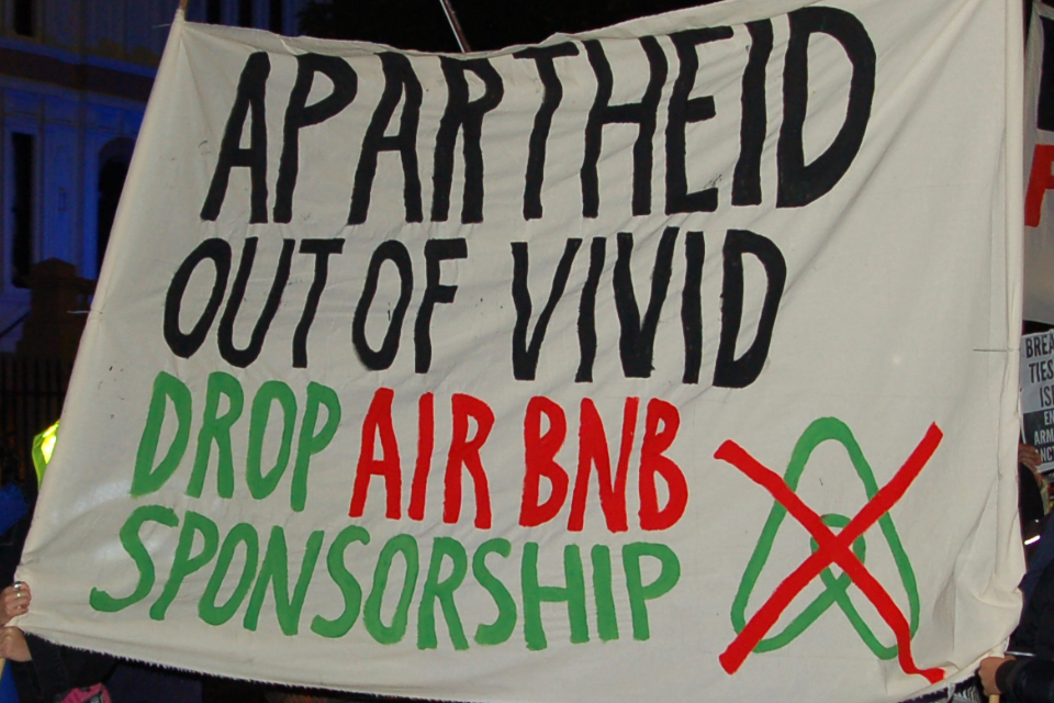 A handmade banner with a crossed-out Airbnb logo reads: Apartheid out of Vivid, drop Airbnb sponsorship