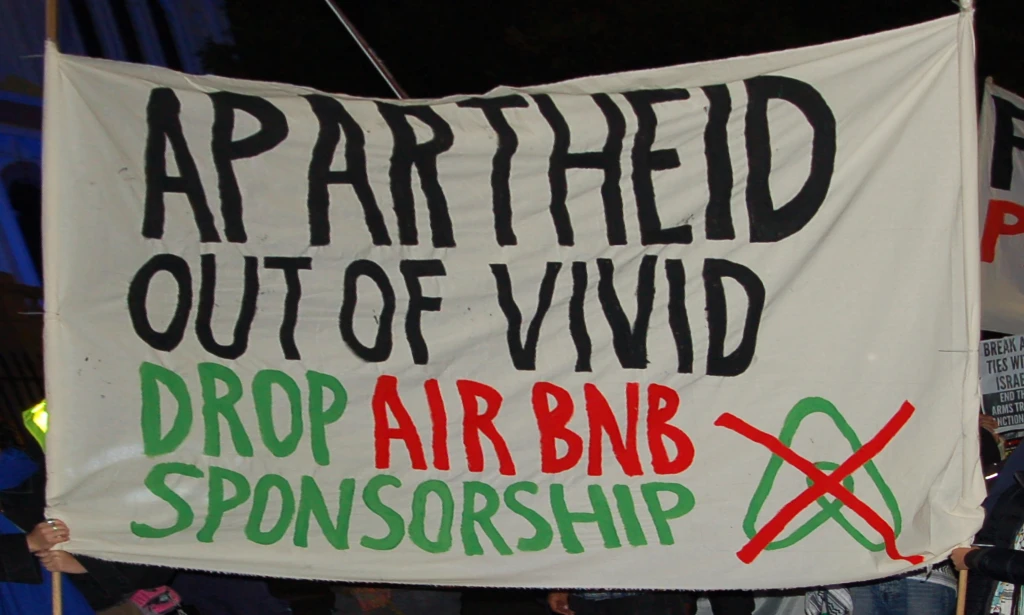A handmade banner with a crossed-out Airbnb logo reads: Apartheid out of Vivid, drop Airbnb sponsorship